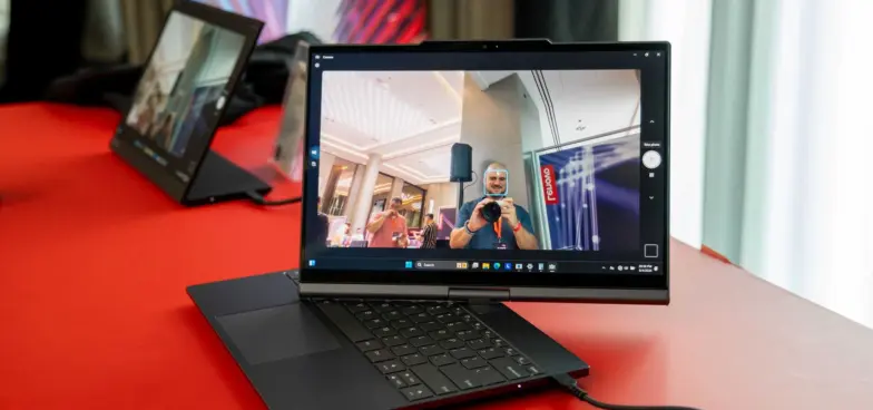 Lenovo laptop with a rotating hinge is displayed on a red surface, showing its screen following the head movements of a user reflected in the screen.